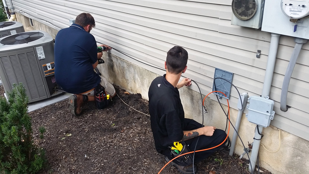 The tech fastens the house cable while a supervisor installs a termination box and ground wire.